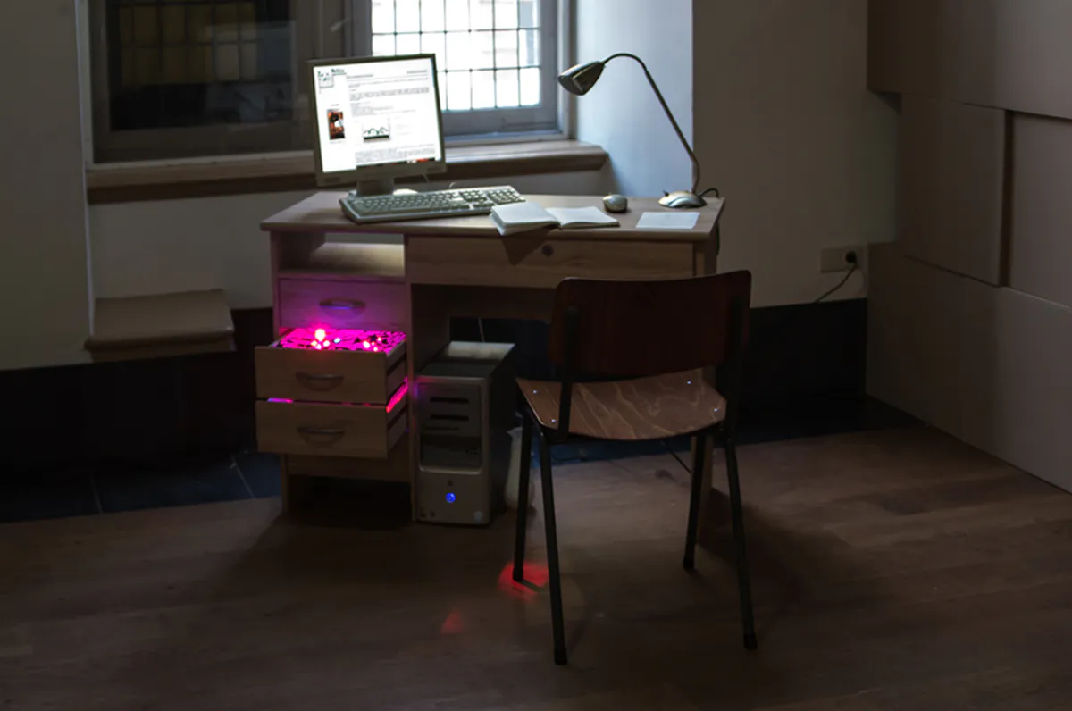 Instalācija ar galdu, datoru, lampu, krēslu un sarkani izgaismotām atvilktnēm izstāžu telpā